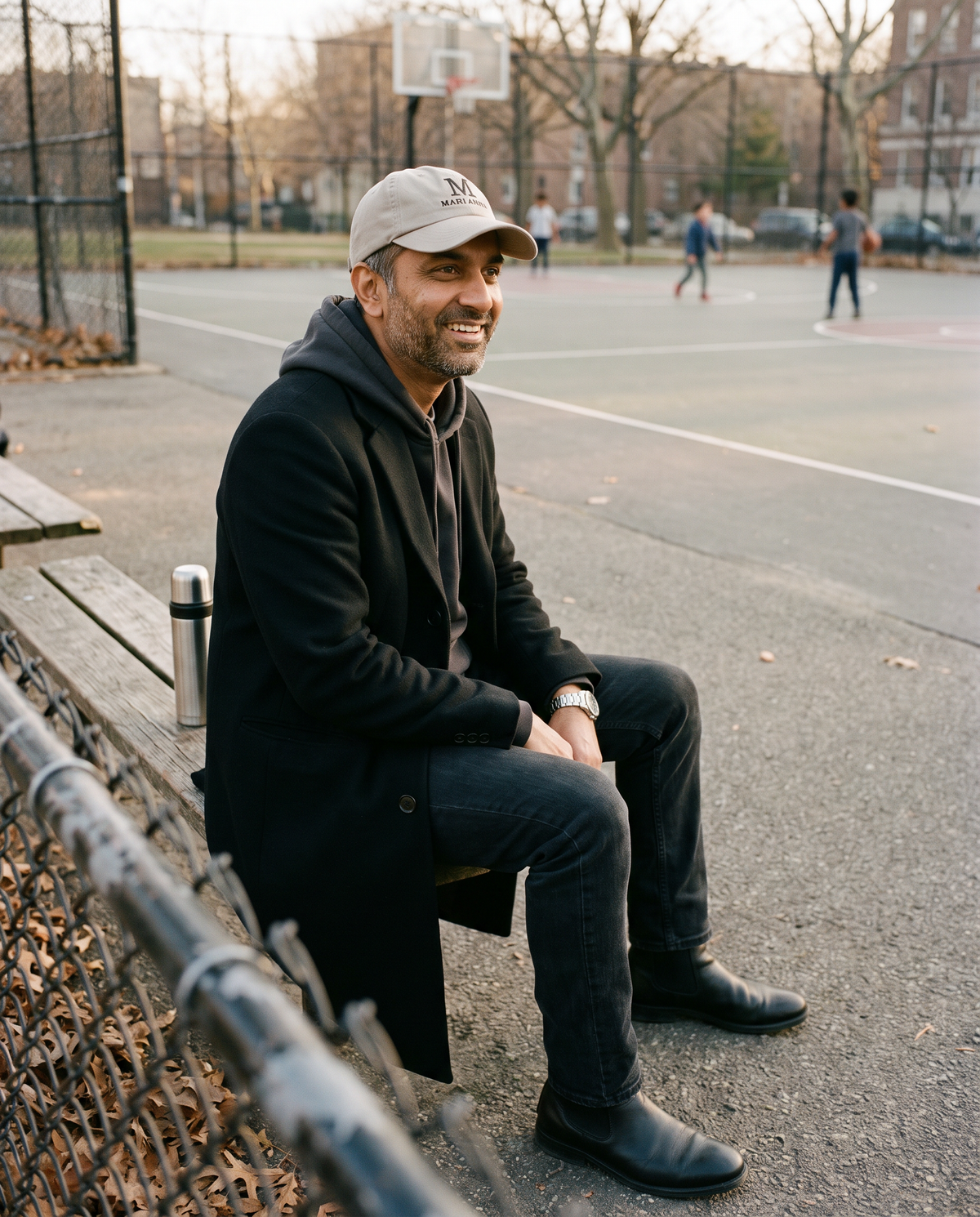 Man in a black overcoat and hoodie on a park bench wearing the stone Mari'Anna dad cap