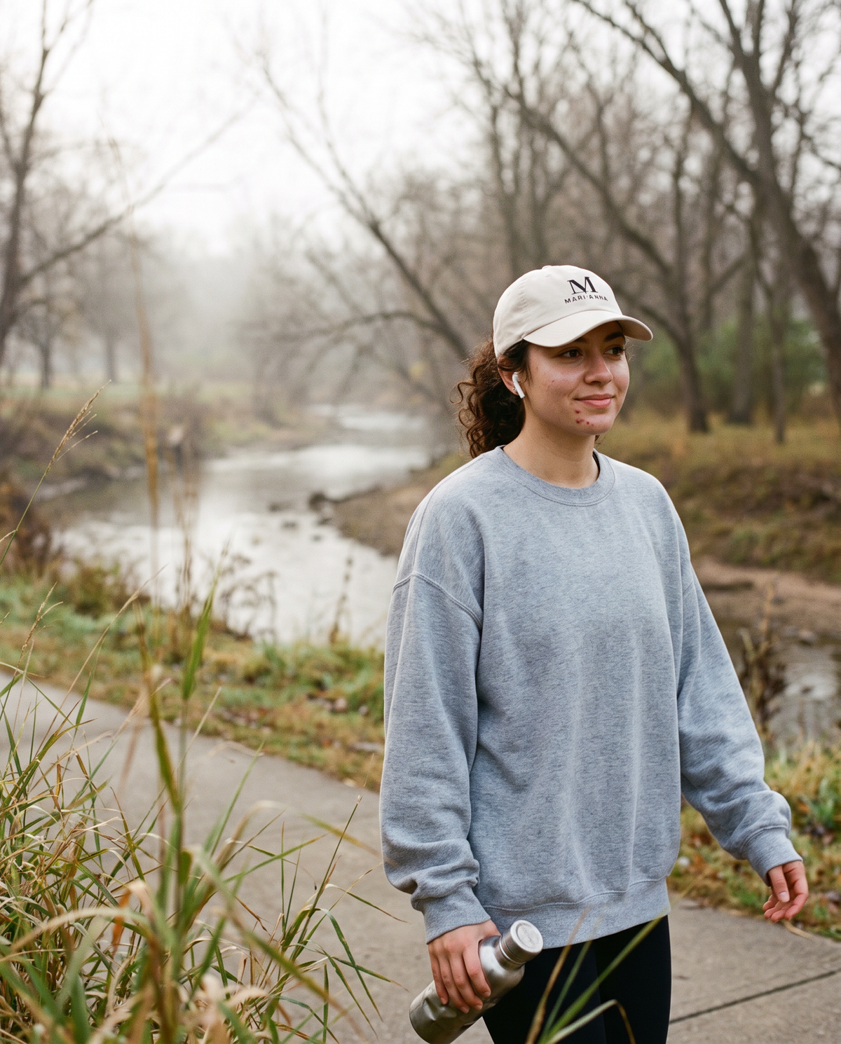 Young woman in a grey sweatshirt on a creek trail wearing the stone Mari'Anna dad cap