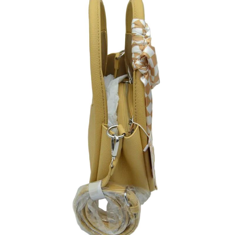 A mustard yellow faux-leather handbag with silver-tone hardware, featuring a matching patterned scarf tied to the handle and a rolled-up strap wrapped in protective plastic.