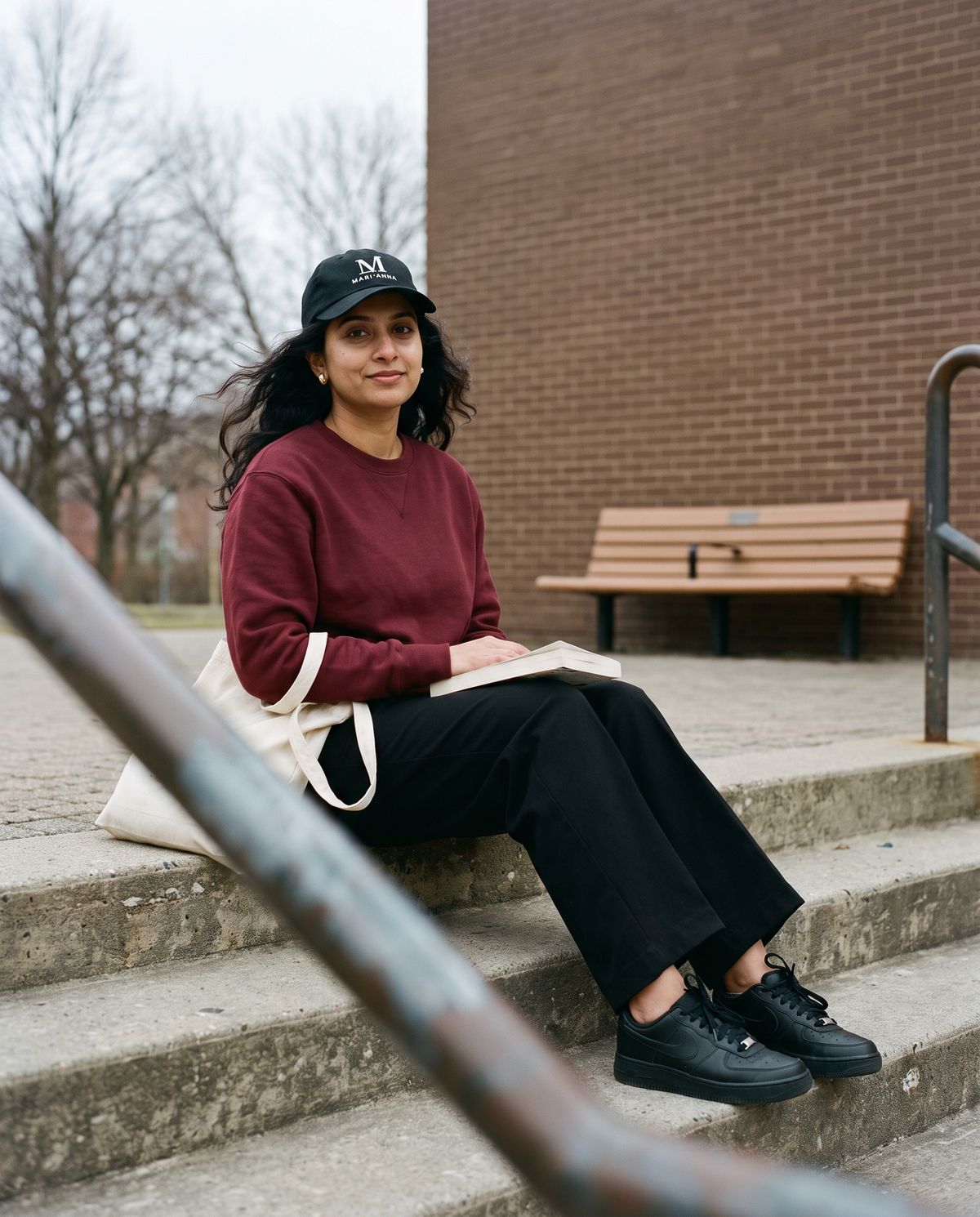 Woman in an oxblood sweatshirt sitting on steps wearing the black Mari'Anna dad cap with the M logo